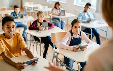 Eine Grundschulklasse mit Tablets für alle Schülerinnen und Schülern, die an einzelnen Tischen sitzen und der Lehrkraft zuhören.