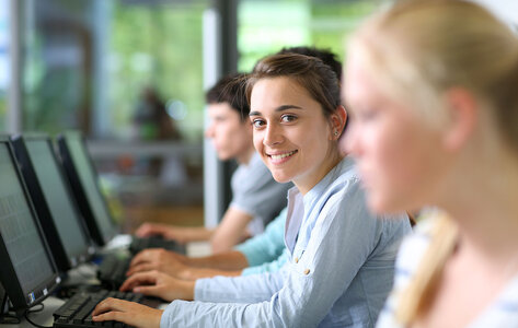 Jugendliches Mädchen sitzt mit anderen Schülerinnen und Schülern in einer Reihe an PCs. Sie schaut lächelnd in die Kamera.