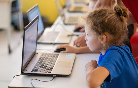 Ein Mädchen in der Grundschule arbeitet hoch konzentriert mit Laptop und Maus. Im Hintergrund sind weitere Schülerinnen und Schüler.
