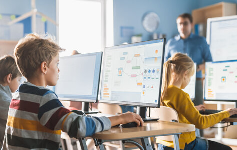 Blick in eine Grundschulklasse: Grundschüler arbeiten am Computer. Auf ihren Bildschirmen sind Flussdiagramme zu sehen. Im Hintergrund beobachtet der Lehrer seine Schülerinnen und Schüler.