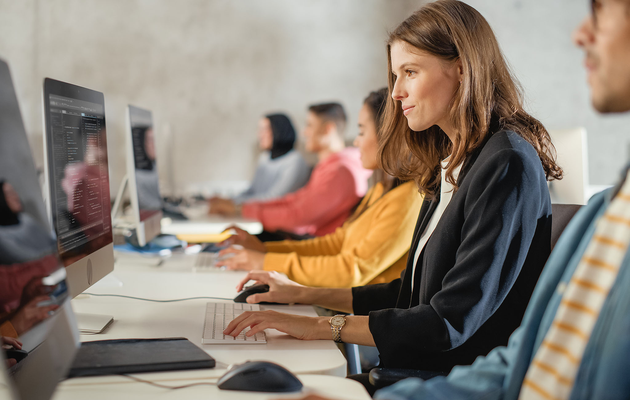 Eine Frau sitzt an einem Computerarbeitsplatz in einem modernen Schulungsraum, mehrere Personen arbeiten im Hintergrund.