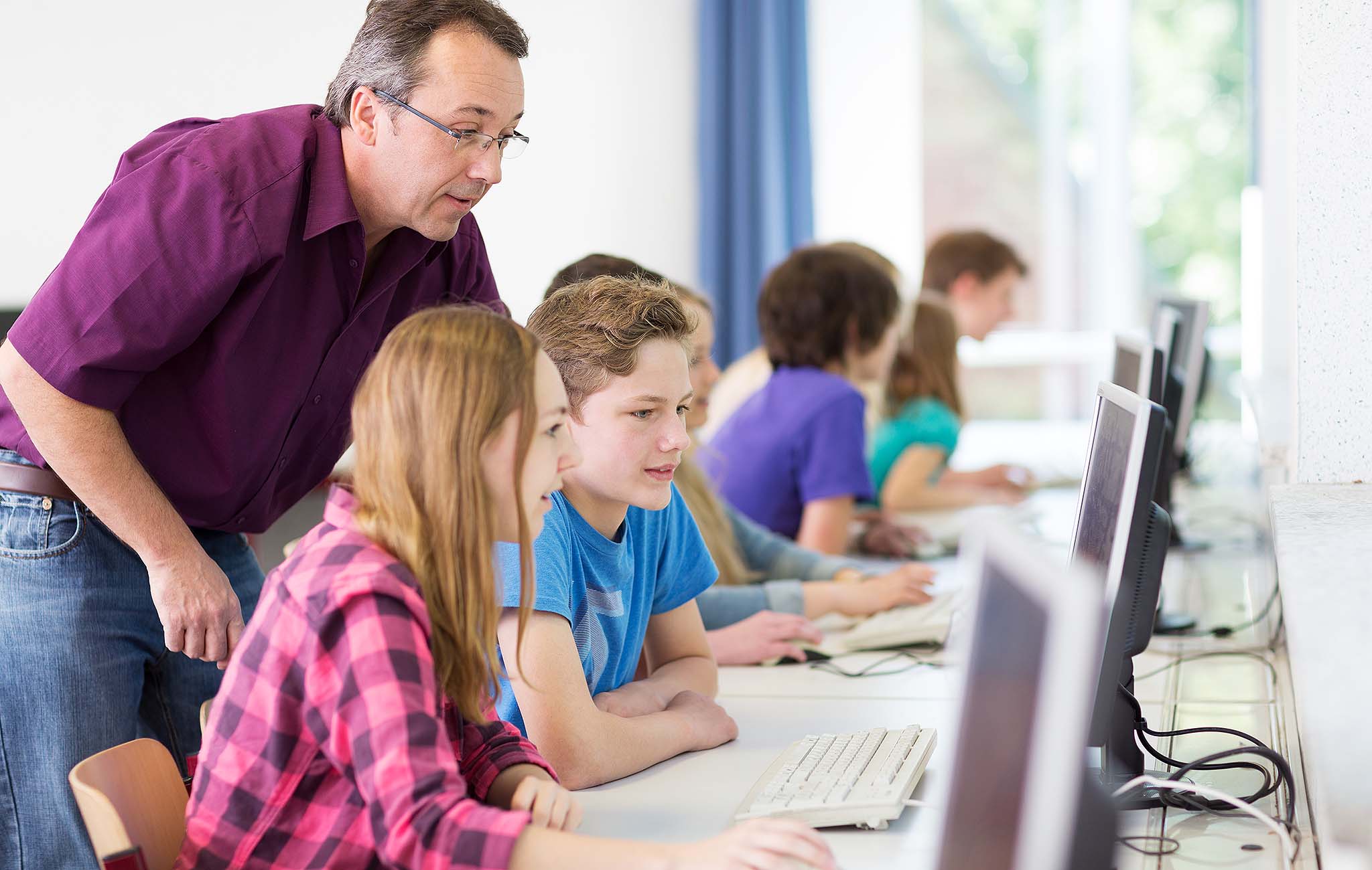 Lehrer unterstützt Schülerinnen und Schüler beim Arbeiten am PC in einem modernen Computerraum – Symbolbild für digitale Bildung mit paedML Linux.