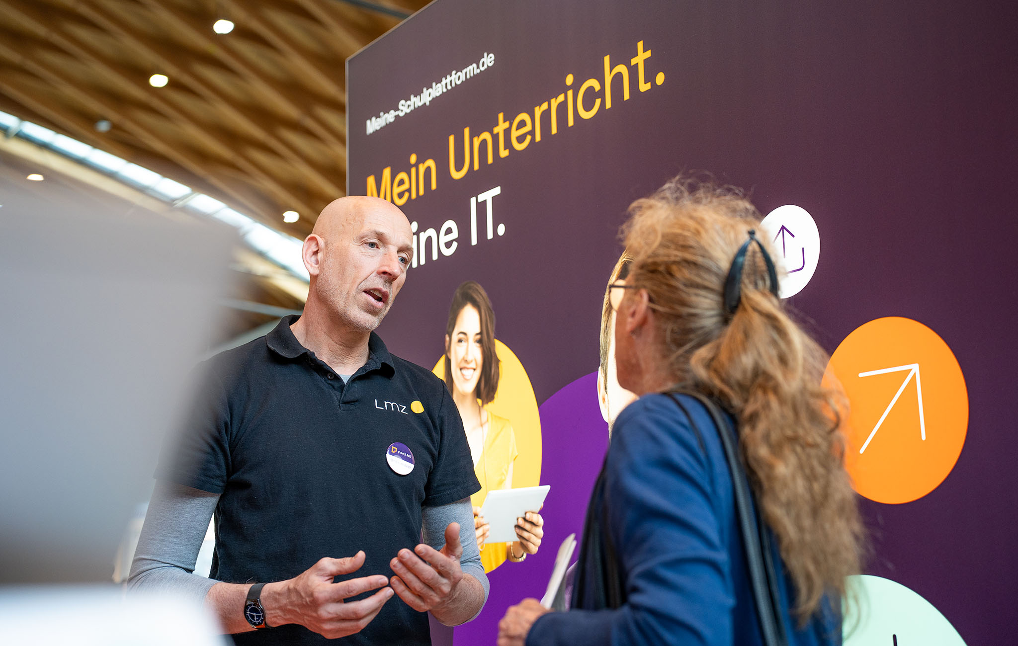 Ein Mann mit dunklem T-Shirt spricht mit einer Frau an einem Messestand. Beide stehen vor einem großen Plakat mit der Aufschrift "Mein Unterricht. Meine IT."