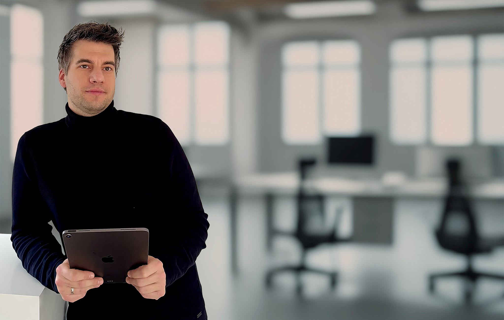 Mann mit Tablet in einem lichtdurchfluteten Büro, steht entspannt und schaut zur Seite – dargestellt im Kontext eines Interviews zur paedML-Lösung.