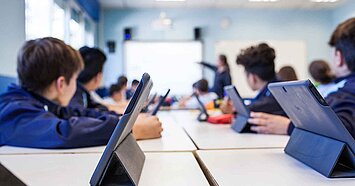 Schüler:innen sitzen im Klassenzimmer an Tischen und arbeiten mit Tablets, die Lehrkraft steht vorn an der Tafel.
