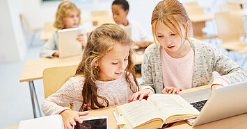 Zwei Grundschülerinnen arbeiten gemeinsam an einem Tisch mit Buch, Tablet und Laptop – Symbolbild für vernetztes Lernen mit paedML in der Grundschule.