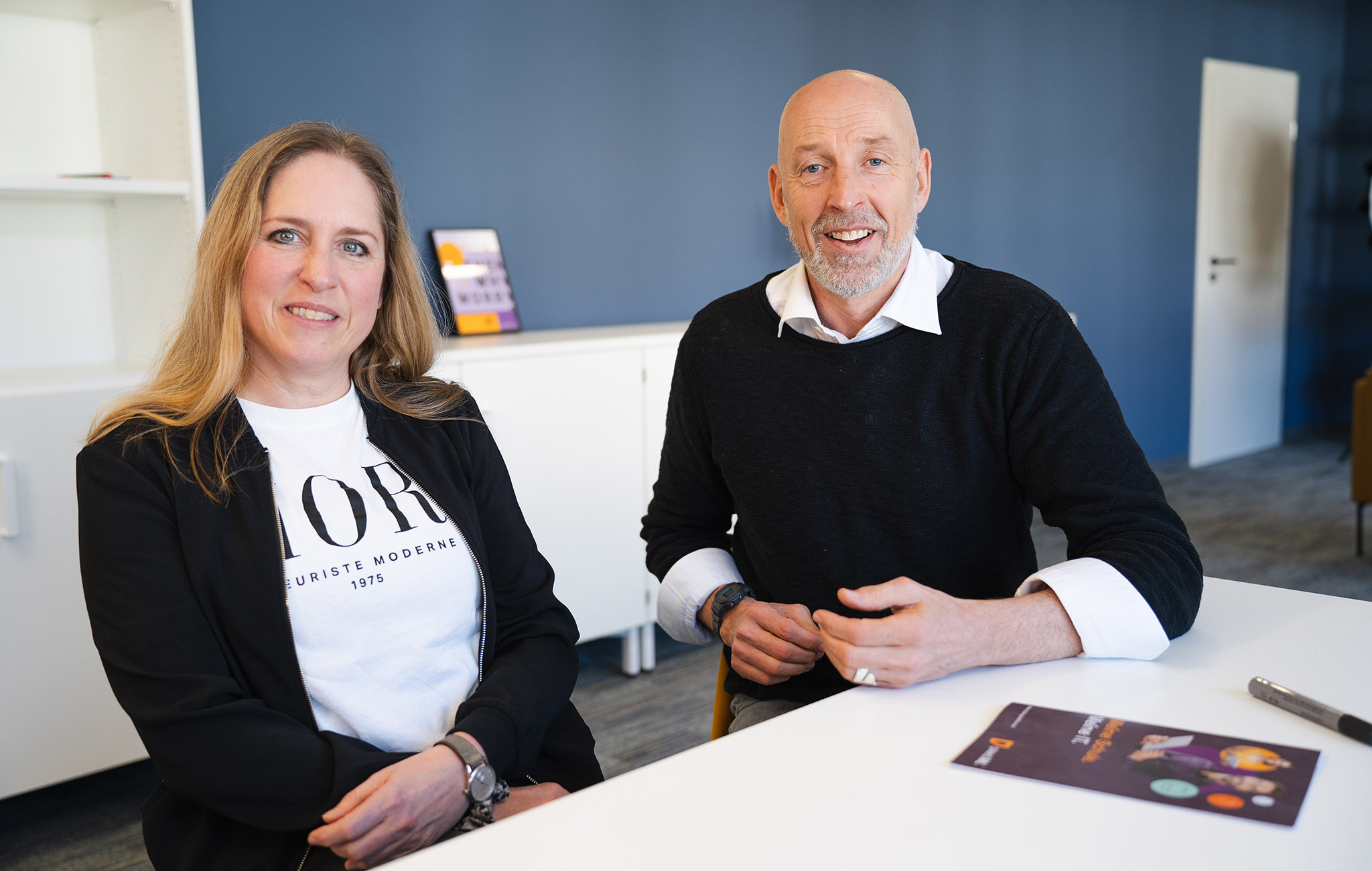 Junge Frau mit blonden langem Haar und Mann sitzen lächelnd an einem weißen Tisch und schauen freundlich in die Kamera.