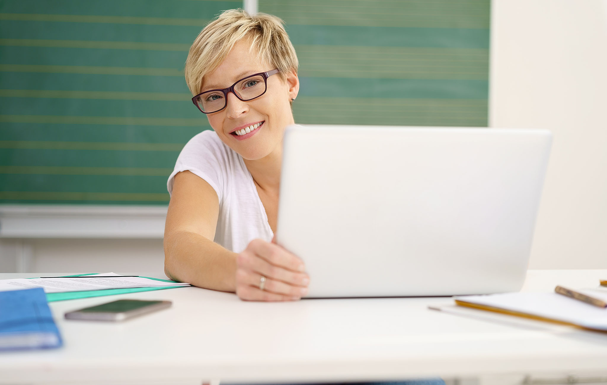 Lehrerin sitzt lächelnd mit Laptop im Klassenzimmer vor der Tafel – Symbolbild für digitale Verwaltung und Benutzerfreundlichkeit mit paedML Linux GS 8.0.
