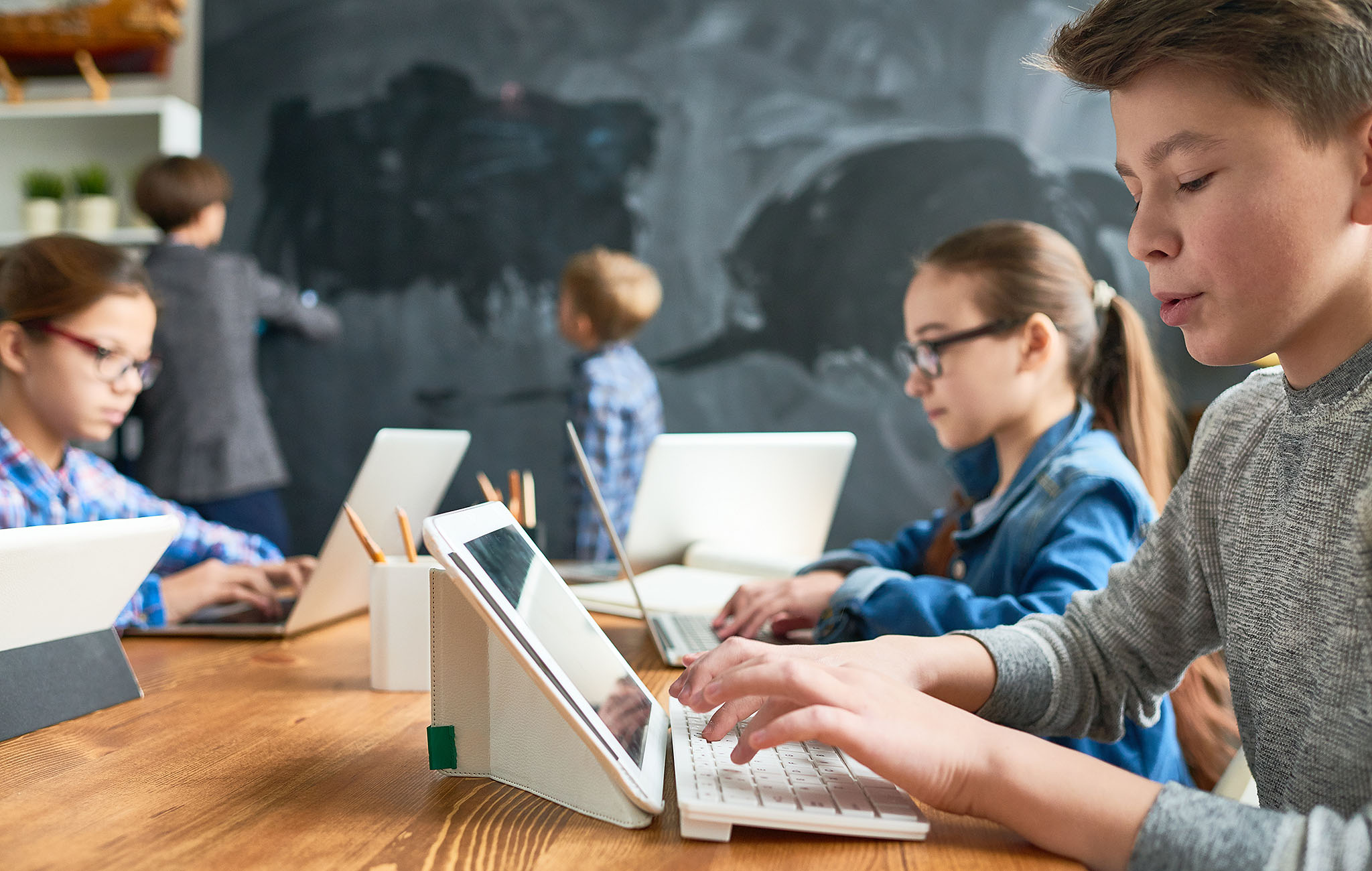 Schülerinnen und Schüler arbeiten mit Tablets und Laptops im Klassenraum, im Hintergrund Kinder an der Tafel – Symbolbild für MDM-Einsatz in der Schul-IT.